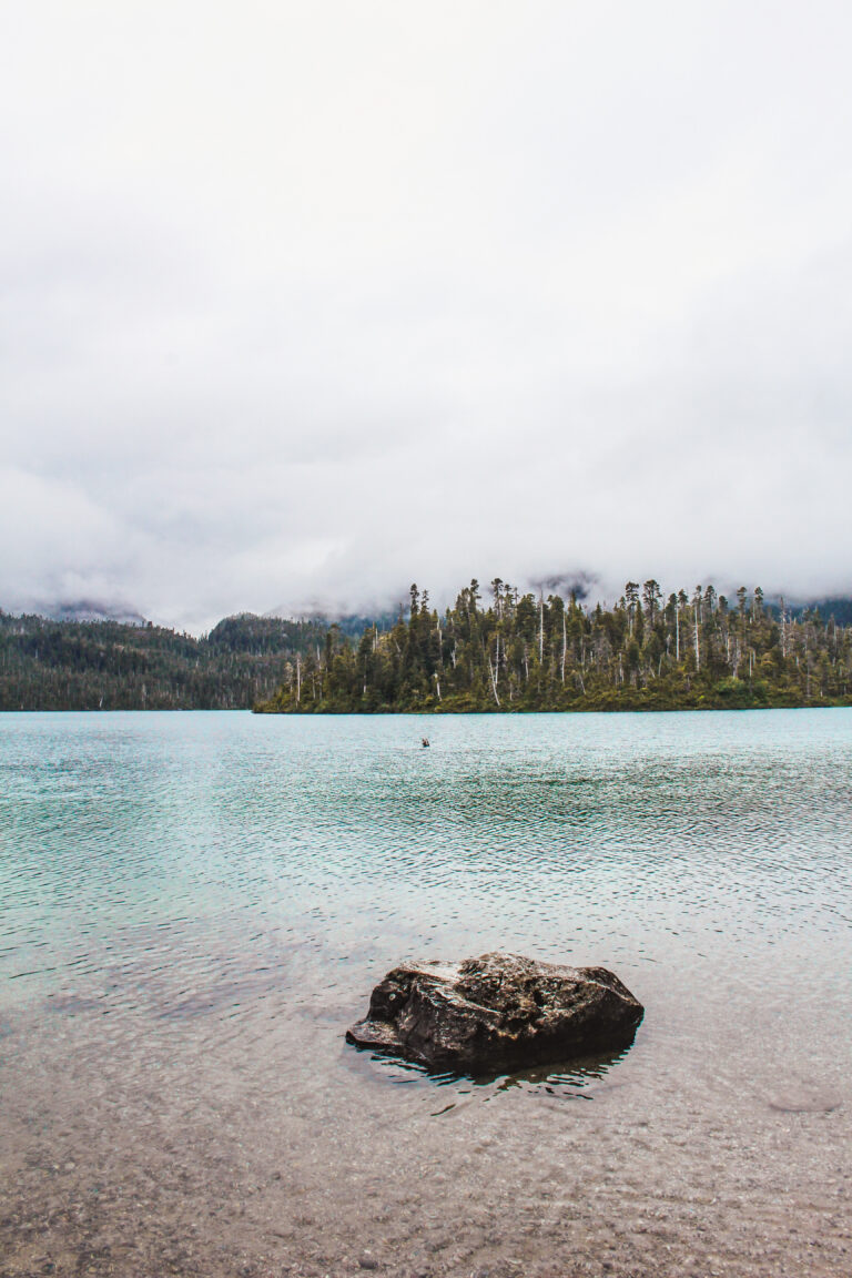 This image is taken from the edge of a lake. There is a medium sized rock protruding from the water in the foreground. Following the rock, the water transitions from brown/translucent (showing the rocks and sand below) to a light blue color. On the other side of the lake are pine trees, and in the distance are mountains shrouded by clouds. This photo was captured on Sitka Island in Alaska, and features the boreal pine tree forests characteristic to the area.