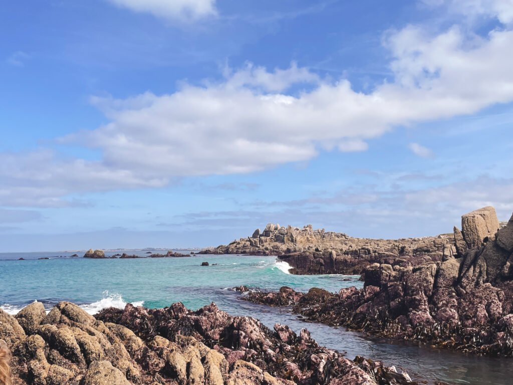 This image is a landscape photo of the seaside. The shoreline is very rocky, zigzagging in and out of frame and cradling the teal ocean, which extends out to the left of the frame. The rocks are a brownish color. It's a sunny day, and the sky is blue with some noticeable fluffy clouds. This image was taken in Brittany, France, on the Atlantic coast.