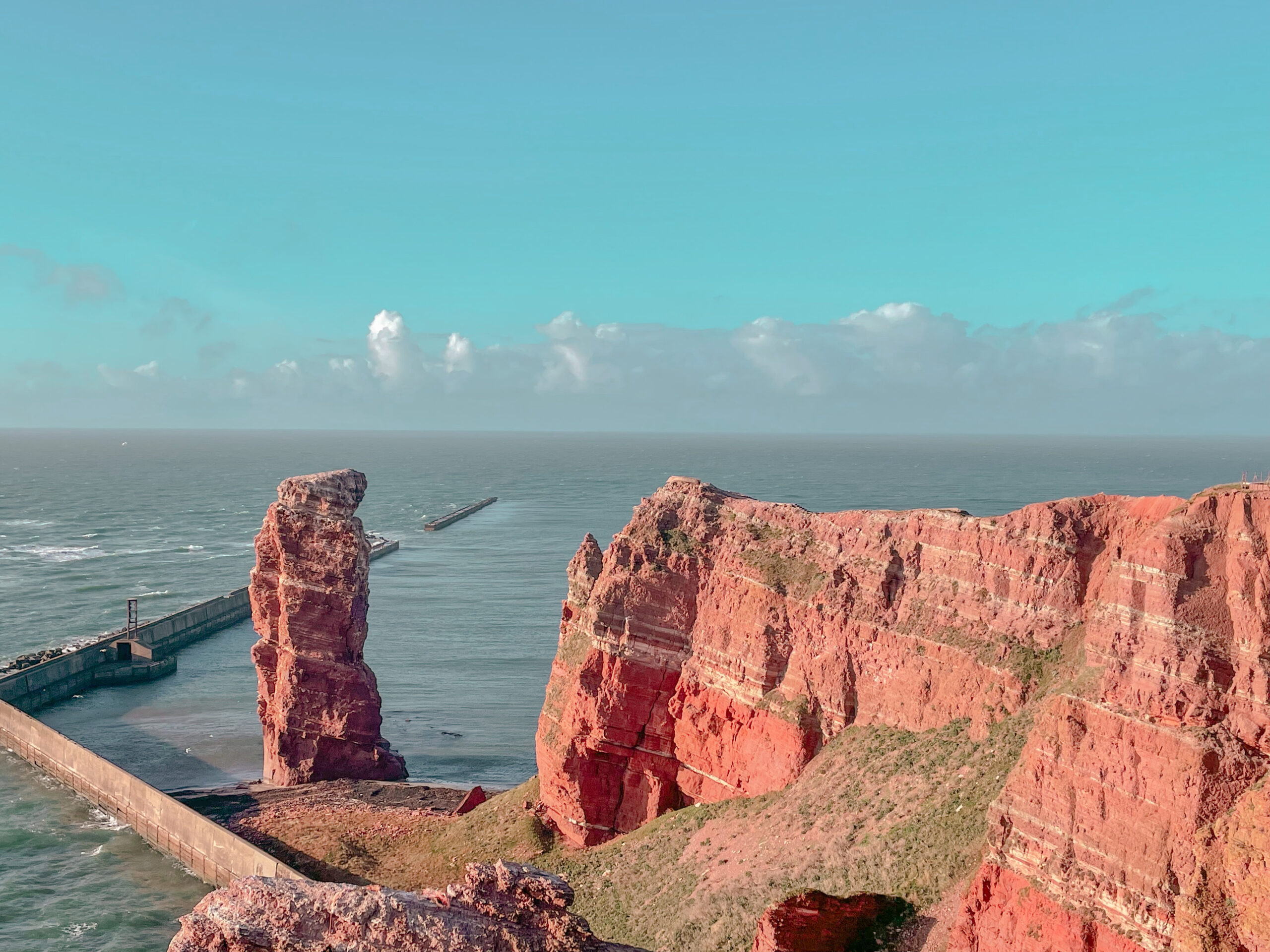 The red cliffs of Helgoland. The image features tall red cliffs in the foreground, the image captured from atop the cliffs nearby. One large rock pillar looms next to the cliffs, reaching a similar height. The sky is clear and blue, with clouds on the horizon, and the sea behind is greyish blue.