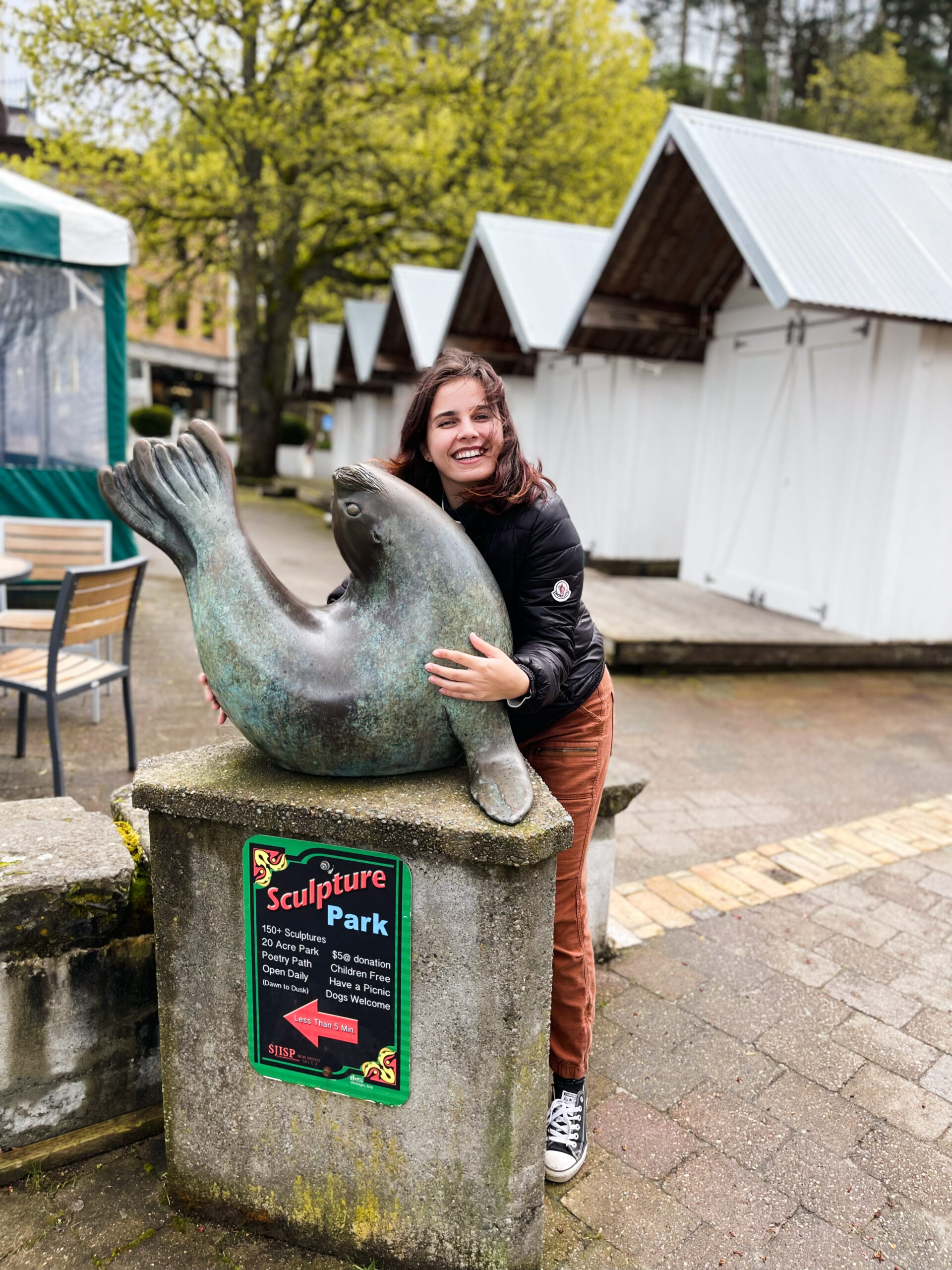 The image features a woman (who is me, LivInSeas, the creator of this website) standing next to a statue of a seal. The seal statue is resting on its belly, with its flippers out for balance. Its tail and head are curled into the air, making it form a seal pose. The woman is standing next to it, cradling it with both arms. She is wearing dark blue converse, tan pants, and a lightweight black puffer coat. Her hair is medium length, just past her shoulders, and is brownish red in color. The wind is blowing it across her face, and she is smiling with her eyes nearly fully shut. The background is not very interesting, some buildings, a brick walkway, and trees.