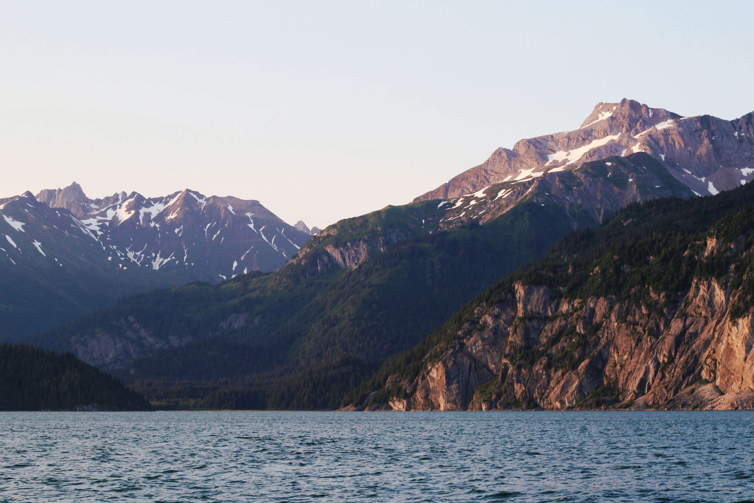 An image captured of mountains rising from the sea in Glacier Bay, Alaska. The sea in the foreground is a soft blue color. The mountains appear to be reflecting the sunset, with a faint pinkish tone. A wall of steep rock rises from the sea on the right side of the screen, covered in green trees on the top. Another mountain rises at a similar angle behind it, with patches of snow at the very top. The bottom is covered in trees, which fade with increased elevation. Behind these two mountains in the distance is a third mountain, broader and with larger patches of snow at the very top. The sky is clear, but is a soft blue/yellow color.