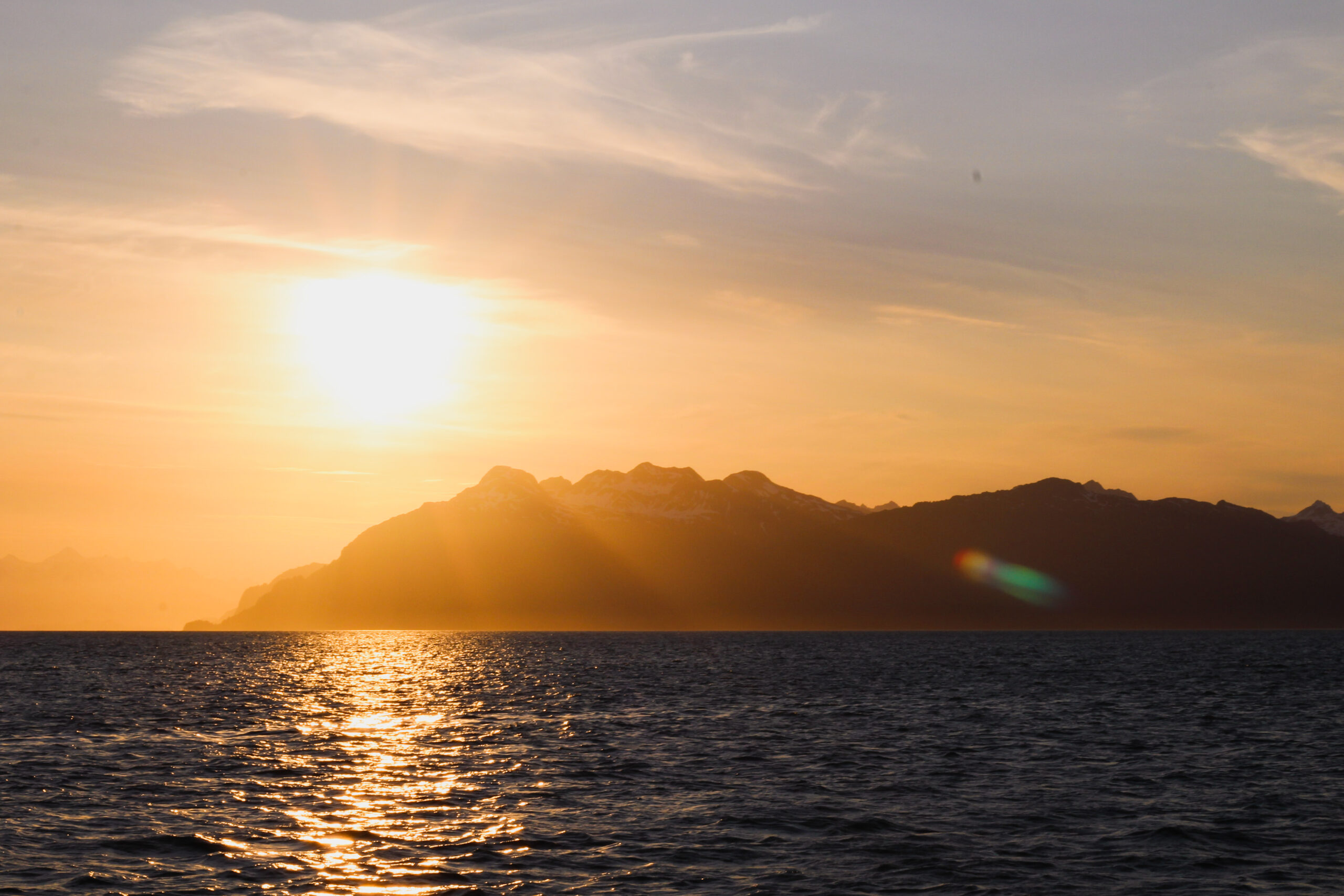 This image was captured on a boat at sunset. The ocean is a dark blue, with a swath of reflected, orange light from the sun. On the horizon is a large mountain, rising from the water in the middle-left of the frame and running off to the right of the screen. The sun is close to skimming the mountains on the left side of the frame, turning the lower part of the sky orange. The upper portion of the sky is a soft, grey color. There is a lens flare in front of the mountain on the right side of the screen.