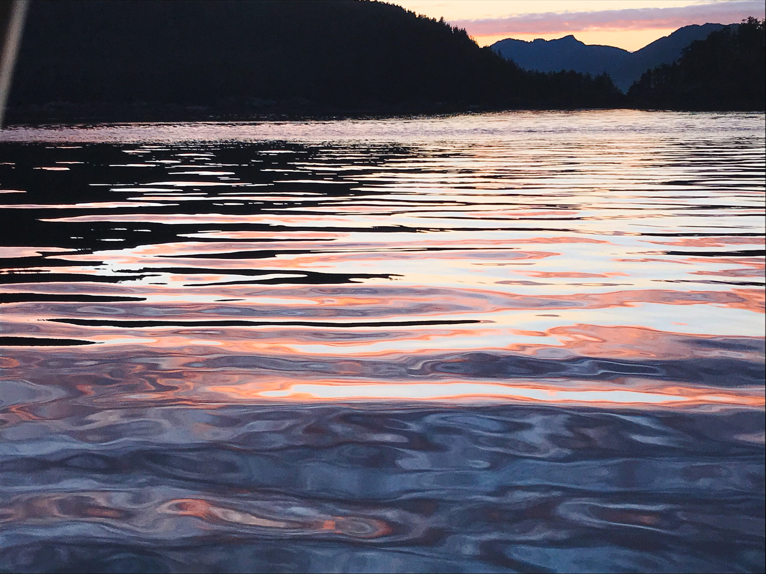 A photograph of water reflecting the sunset sky. The rippling sea surface is the focus of the photo, but hills rise from the sea in the background. The water is a purpleish blue in some parts, but reflects the black of the hills in some parts and the white/orange/pink of the sunset sky in other parts.