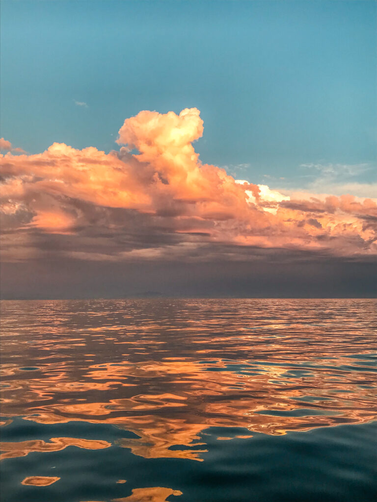 An image taken of the sea at sunset. The ocean is dark blue in the very foreground, but reflects the orange and dark grey of the sky and clouds from the foreground to the midground. At the middle of the photo, the sea ends and cloudy sky begins, switching from grey to puffy, tall formations of orange and yellow clouds. The sky behind the clouds is blue.