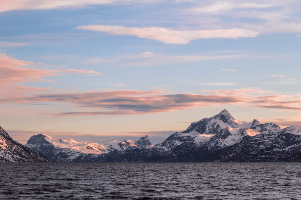Snow-covered mountains looming over a dark grey ocean at sunset. The mountains are in the distance and swathed by pinkish light from the sunset, with pink-grey clouds drifting above them. The sky transitions from yellow to blue. Image was taken from a boat (not visible) on the sea in Nuup Kangerlua Fjord, Greenland.