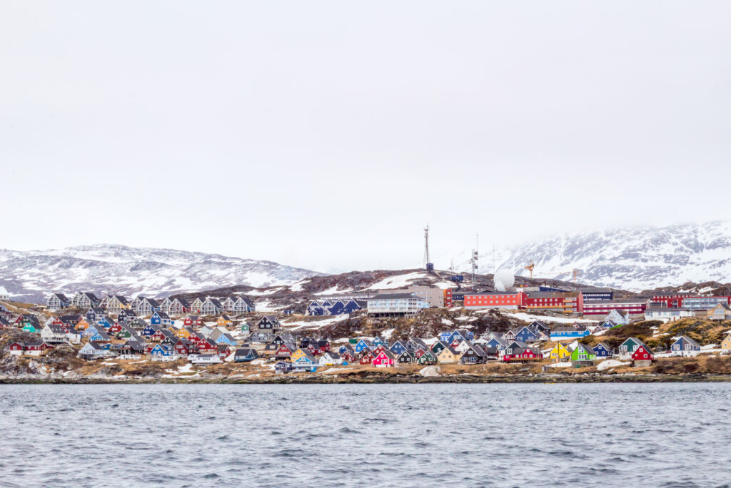 The colorful houses of Nuuk, captured from the sea. The houses all look like colorful jewels situated on a rocky, brown shoreline. Many homes and buildings in all shades of the rainbow sprawl from the left to the right side of the frame. The ocean in the front of the photo is grey, and it is a cloudy day. In the background are two short, sloping snow-covered mountains.