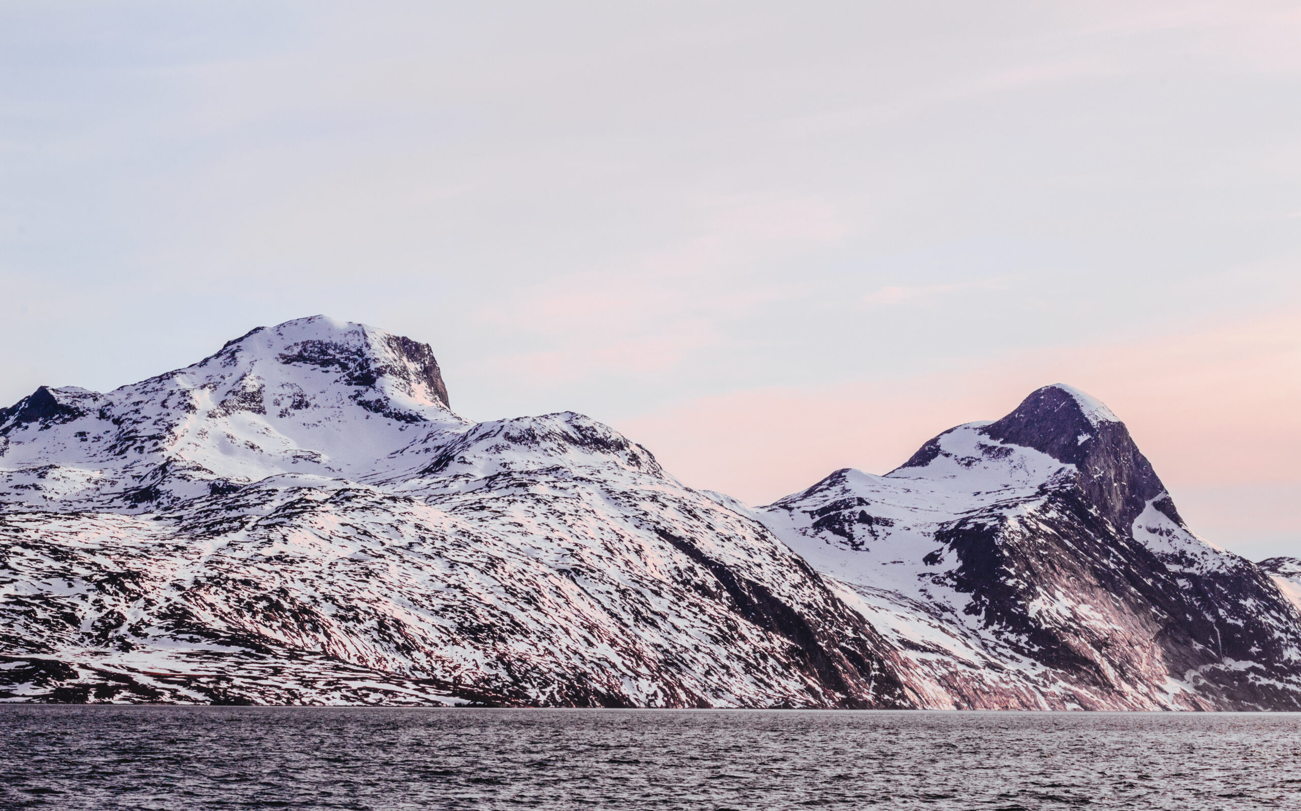 A landscape photo of snowy mountains next to the ocean. This photo was captured by LivInSeas in the Nuup Kangerlua fjord in Greenland. It's sunset, and the sky is hazy with soft blue and fuzzy pink clouds. The mountains and snow are reflecting a slight pinkish hue as well. There are two main mountains visible, one on the left and one on the right side of the screen. The ocean sits in the foreground.